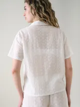 Camisa de manga corta para dormir confeccionada en broderie de algodón, con cuello solapa y cierre frontal con botones. Presenta un bolsillo aplicado en el pecho.