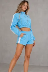 Conjunto deportivo de dos piezas color celeste con franjas blancas en los laterales. Incluye una campera crop de manga larga con cierre frontal, bolsillos y detalles de pedrería brillante en los hombros, y un short a juego con cintura elástica y bolsillos.
