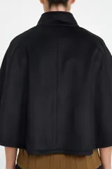 Chaqueta tipo capa de color negro, con cuello redondo y cierre frontal mediante botones grandes. Presenta un diseño minimalista y elegante con corte holgado.