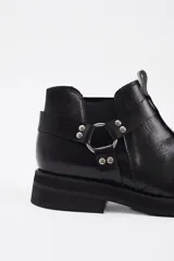 Botineta de cuero vacuno color negro, con diseño estilo western que incluye tiras decorativas, argollas metálicas y tachas plateadas. Cuenta con suela de goma y taco bajo cuadrado.