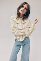Blusa de viscosa color beige con estampado de lunares negros. Presenta cuello con volados, cordón fino para ajustar, botonadura frontal y mangas largas con puños elásticos fruncidos.