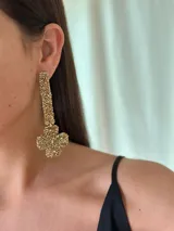 Aro colgante de diseño geométrico compuesto por una barra vertical y una pieza inferior en forma de cruz, ambos totalmente recubiertos por mostacillas en tonos dorados y metalizados.