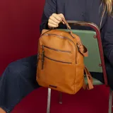 Mochila de cuero sintético con textura granulada, color verde oliva. Cuenta con dos bolsillos frontales con cierre metálico, asa superior rígida y correas ajustables de cinta textil. Incluye placa metálica decorativa en la parte inferior.