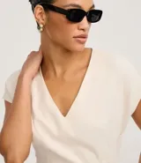 Enterito corto de mujer color beige claro, con escote en V, mangas cortas tipo capa, trabillas en la cintura y pliegues frontales. Incluye un cinturón fino trenzado de cuerda.