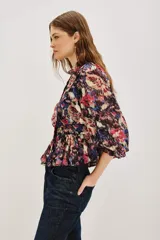 Camisa de manga larga con estampado floral y detalles en hilo metalizado. Presenta cuello alto con volados, cierre frontal con botones y puños con volados. El diseño incluye un corte entallado en la cintura con un ligero peplum.