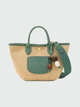 Cartera tipo crossbody de la línea Le Panier Pliage de Longchamp, confeccionada en paja trenzada con detalles en cuero blanco. Presenta un diseño pequeño con asas superiores, correa larga ajustable y extraíble, y un parche frontal con el logo de la marca. Incluye un adorno colgante con cuentas de madera.