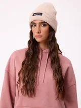 Gorro tipo beanie de punto acanalado en color beige, con parche rectangular negro con el logo de la marca en el frente.