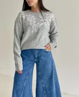 Sweater de punto gris con cuello redondo, manga larga y detalle de bordado floral en color blanco en la parte superior delantera.