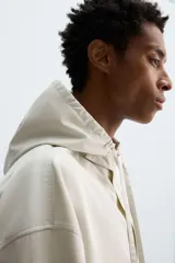 Sobrecamisa de corte oversized confeccionada en tejido compacto de algodón. Presenta cuello subido con capucha ajustable mediante cordones, cierre frontal con botonadura, manga larga con puños abotonados y dos bolsillos de plastrón en la parte frontal.