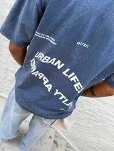 Remera oversize de algodón con efecto lavado, color azul oscuro. Presenta un estampado minimalista en blanco en el pecho con texto en dos líneas: "URBAN LIFESTYL" y "RAQRA YTIJAU" (lectura invertida de "QUALITY APPAREL"), y la palabra "OSTEN" en el lado derecho.