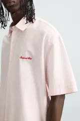 Camisa de algodón color rosa pálido, corte boxy fit, con cuello solapa, manga corta y abotonadura frontal. Presenta un texto bordado en contraste de color rojo en el delantero.