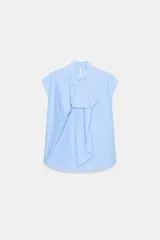 Camisa sin mangas confeccionada en algodón con estampado de rayas verticales en tonos celeste y blanco. Presenta cuello redondo con detalle de lazada frontal, corte holgado y bajo redondeado asimétrico. Cierre posterior con abertura y botones.