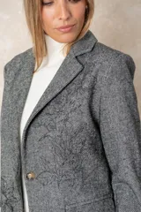 Blazer de corte sastre con diseño de espiga en tonos grises. Presenta un bordado floral lineal en el lateral, solapas clásicas, cierre frontal con un botón y bolsillos con solapa.