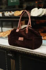 Bolso tipo duffle color verde militar, confeccionado en gamuza, con manijas de cuero marrón. Incluye bolsillos laterales, uno con clip, cierre posterior y un bolsillo interno amplio. Detalle de bandera y logo bordado en los laterales.