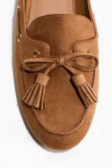 Mules de gamuza color marrón tostado, estilo mocasín, con puntera redondeada, detalle de lazo con borlas en el empeine y diseño destalonado.