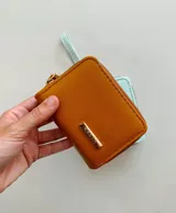 Billetera compacta de cuero sintético color tostado con cierre perimetral y placa metálica dorada con el logo de la marca en el frente. Incluye una correa de mano desmontable.
