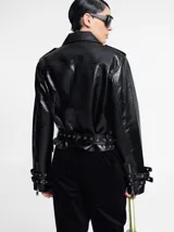 Chaqueta biker negra de cuero sintético con tachas plateadas en los hombros y solapas, cierre asimétrico con cremallera, cinturón ajustable con hebilla y detalles metálicos.