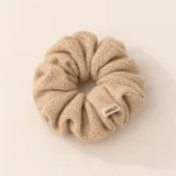 Colero de pelo tipo scrunchie con textura suave y abrigada, en color beige. Presenta un diseño voluminoso con una etiqueta pequeña de la marca Amalfi.