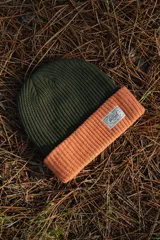 Gorro de lana tejido a punto, color verde oliva en la parte superior y naranja en el borde inferior. Tiene una etiqueta rectangular cosida en el frente con el logo de la marca Denali.