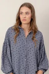 Camisa de corte oversize y diseño holgado, color celeste con estampado de pequeños corazones negros. Presenta cuello clásico, cierre frontal con botones y mangas largas abullonadas con puños ajustados.