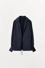 Camisa de manga larga color azul marino, confeccionada en tejido satinado. Presenta cuello solapa, puños anchos y cierre frontal mediante lazada ajustable en el centro.
