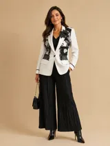 Blazer entallado color blanco con sutiles brillos, intervenido artesanalmente con apliques de texturas y encaje negro en las solapas, incluyendo un detalle floral decorativo.