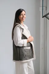 Chaleco de gamuza sintética en color verde oliva con interior y bordes de corderito beige. Presenta solapas amplias, dos bolsillos frontales aplicados y un diseño abierto sin cierre.
