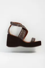 Sandalias de cuero color marrón con plataforma y taco de cuña. Presentan tiras cruzadas con apliques de tachas y hebilla de ajuste en el tobillo.