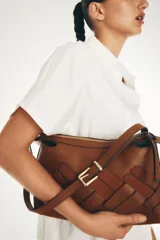 Cartera marrón con diseño tejido en la parte frontal, cierre superior y correa ajustable con hebilla metálica.
