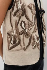 Chaleco de lana color beige con bordados florales artesanales en tonos marrones y blancos. Presenta un diseño sin mangas con volados en los hombros y cierre frontal mediante lazos negros ajustables.