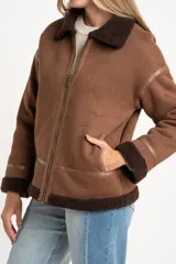 Chaqueta tipo gamulán de calce oversize en color marrón, con cuello y ribetes de corderito en contraste. Presenta cierre frontal de cremallera, bolsillos laterales y detalles de tiras decorativas en material sintético.