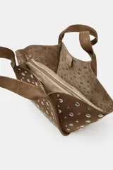 Bolso tipo shopper de hombro, confeccionado en gamuza color marrón, decorado con tachas metálicas plateadas en toda su superficie. Cuenta con doble asa de hombro y cierre mediante botón imantado.