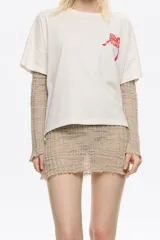 Camiseta de algodón color blanco hueso, de corte boxy y holgado, con escote redondo y mangas cortas. Presenta un pequeño logo en relieve de color rojo en el área del pecho.