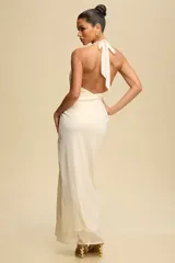 Vestido largo de fiesta color crema, con diseño drapeado, escote halter y espalda descubierta. Presenta un detalle metálico dorado en el hombro y una abertura frontal en la falda.