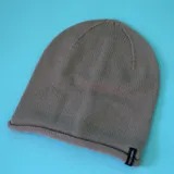 Gorro tipo beanie de tejido de punto, color marrón claro, con borde enrollado y etiqueta de marca en el lateral.