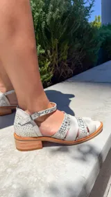 Sandalia plana de cuero, color blanco y plateado, con tiras anchas en el empeine con textura de cocodrilo y una tira fina ajustable en el tobillo. Posee un pequeño tacón de madera.