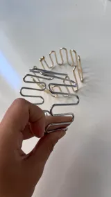 Brazalete abierto de metal con diseño ondulado o en forma de 'S' repetida. Se observa en dos acabados: plateado y dorado.