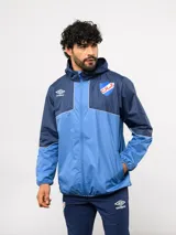 Campera de lluvia Umbro del Club Nacional de Football, con diseño de bloques de color en rojo y azul marino. Presenta capucha, cierre frontal, puños elásticos y detalles de piping blanco. Incluye logo de Umbro y escudo del club en el pecho.
