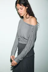 Camiseta de cuello barco y manga larga tipo murciélago, confeccionada en tejido de punto gris melange. Presenta detalle de tachas metálicas a lo largo de los hombros y las mangas.