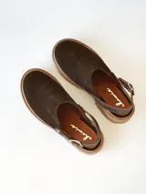 Zueco de cuero gamuzado color chocolate, con diseño de punta cerrada y tira ajustable con hebilla metálica en el talón. Presenta una suela de plataforma dentada en tono marrón claro.