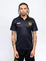 Remera polo de poliéster color negro con cuello clásico y tapeta de dos botones. Presenta el escudo del Club Atlético Peñarol bordado en el pecho y el año de fundación (1891) estampado en blanco. Tiene detalles en amarillo en los hombros y el escudo del club repetido en la parte inferior.