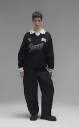 Sweater de punto negro con cuello polo en color crema, manga larga y diseño oversize. Presenta bordados en el frente con la inscripción 'Ravers', el número '90' y letras cursivas, con terminaciones en punto rib en puños y cuello.