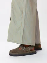 Slipper tipo guillermina confeccionada en gamuza color marrón, con plataforma de goma negra y doble hebilla ajustable. Presenta un pespunte decorativo en el borde inferior y cuenta con interior recubierto en cuero.