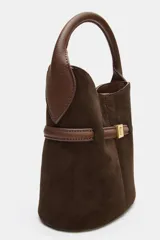 Bolso tipo saca de tamaño mini, confeccionado en gamuza color marrón oscuro con detalles en cuero marrón. Presenta un asa de mano fija, una tira decorativa horizontal con hebilla metálica dorada en el frente y costuras verticales visibles.