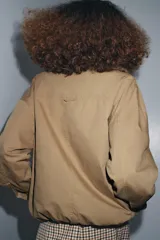 Chaqueta tipo bomber de color beige, con cuello solapa y aplicación extraíble de pelo sintético. Presenta cierre frontal con botones a presión ocultos, bolsillos delanteros y bajo con elástico.