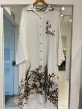 Vestido largo estilo camisero color blanco, con cierre frontal de botones, bolsillos superiores y un gran estampado floral en tonos marrones y verdes en la parte inferior y en el bolsillo.