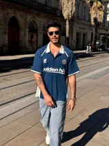 Camiseta tipo polo de manga corta con calce holgado, confeccionada en tejido técnico. Presenta cuello camisero en color blanco, detalles en contraste en hombros y mangas, y estampados gráficos en el frente con el logo de la marca y la inscripción golden hill.