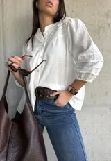Camisa blanca de estilo romántico con cuello mao, botones frontales y mangas tres cuartos abullonadas con detalle de frunces. Presenta un diseño con textura sutil y costuras decorativas en la parte superior delantera.