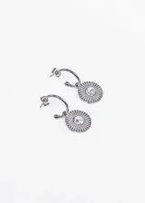 Aros colgantes de acero quirúrgico plateado con dije circular con diseño de rayos y piedra blanca en el centro.