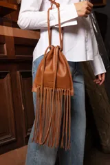 Cartera tipo morral color tostado, de diseño tipo saco con cierre de cordón ajustable y tiras largas. Cuenta con flecos largos colgando en la base y correa larga desmontable para cruzar.